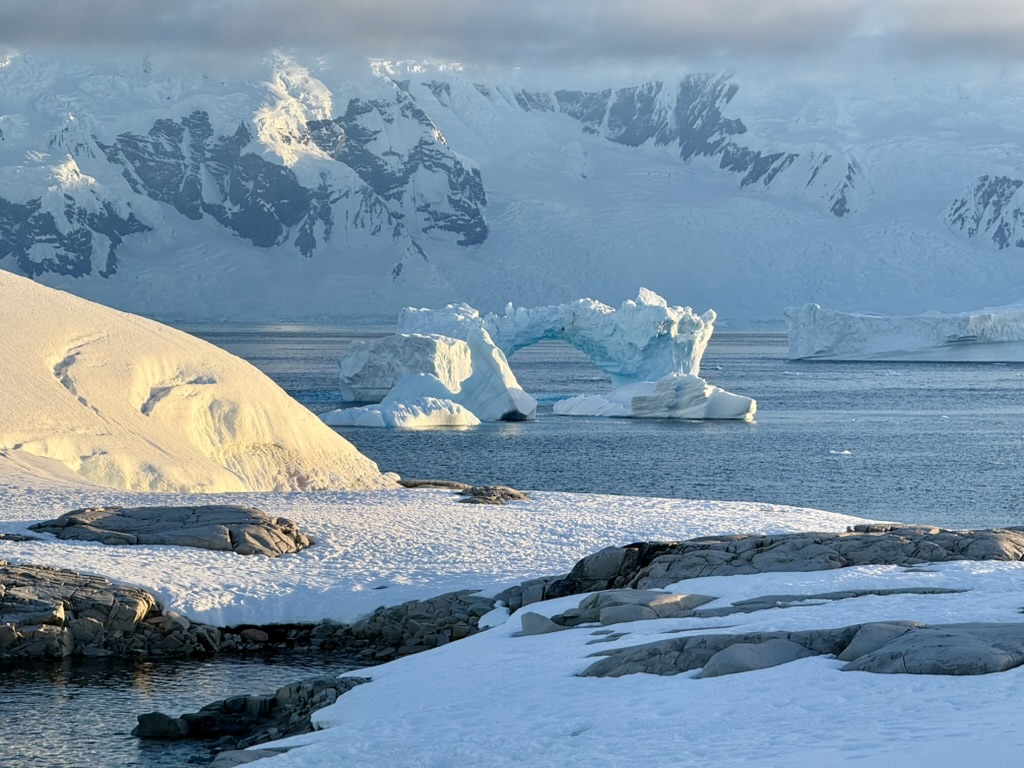 Warm low-angle light across Antarctic snow, an ice arch and immense mountain walls